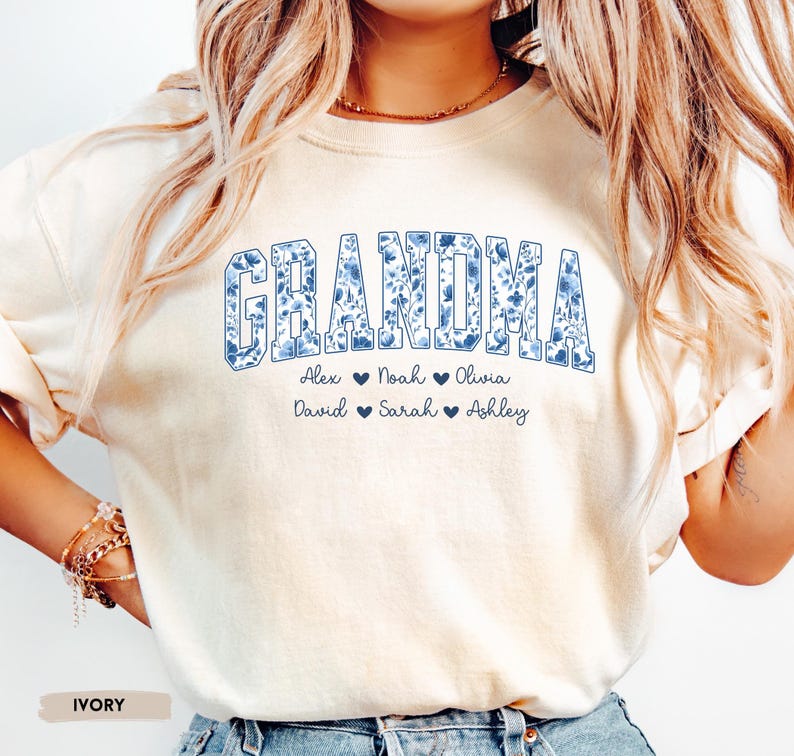 May include: Cream-coloured sweatshirt featuring the word "GRANDMA" in a blue floral design. Underneath are the names Alex, Noah, Olivia, David, Sarah, and Ashley, each with a heart symbol. The word "IVORY" is printed at the bottom.