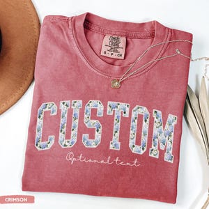 May include: Crimson-colored t-shirt with the word "CUSTOM" in large, floral-patterned letters. Below the word "CUSTOM" is the text "Optional text". A silver necklace with a pendant rests on the shirt. The shirt is labeled "Comfort Colors".