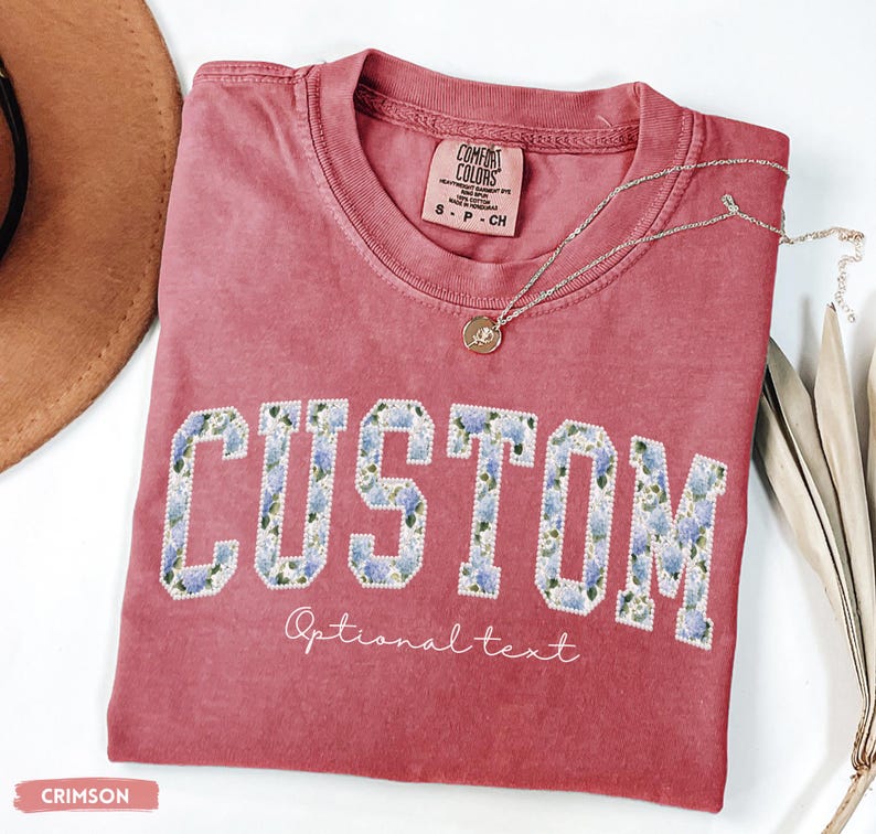 May include: Crimson-colored t-shirt with the word "CUSTOM" in large, floral-patterned letters. Below the word "CUSTOM" is the text "Optional text". A silver necklace with a pendant rests on the shirt. The shirt is labeled "Comfort Colors".