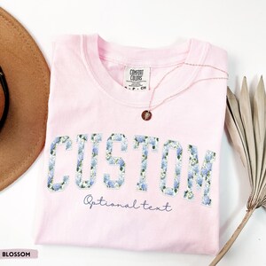May include: A light pink t-shirt with the word "CUSTOM" in large, blue floral letters. Below the word "CUSTOM" is the text "Optional text" in a script font. A gold necklace with a pendant rests on the shirt.
