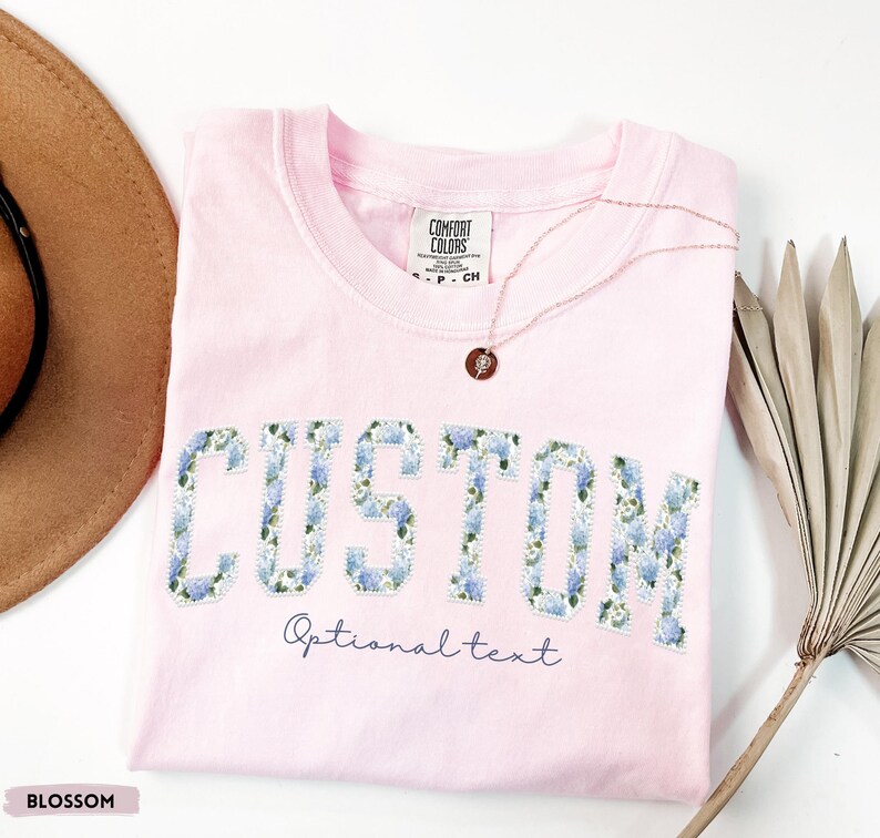 May include: A light pink t-shirt with the word "CUSTOM" in large, blue floral letters. Below the word "CUSTOM" is the text "Optional text" in a script font. A gold necklace with a pendant rests on the shirt.