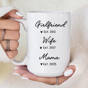 May include: White ceramic mug with black text that reads "Girlfriend Est. 2011", "Wife Est. 2017", and "Mama Est. 2025".