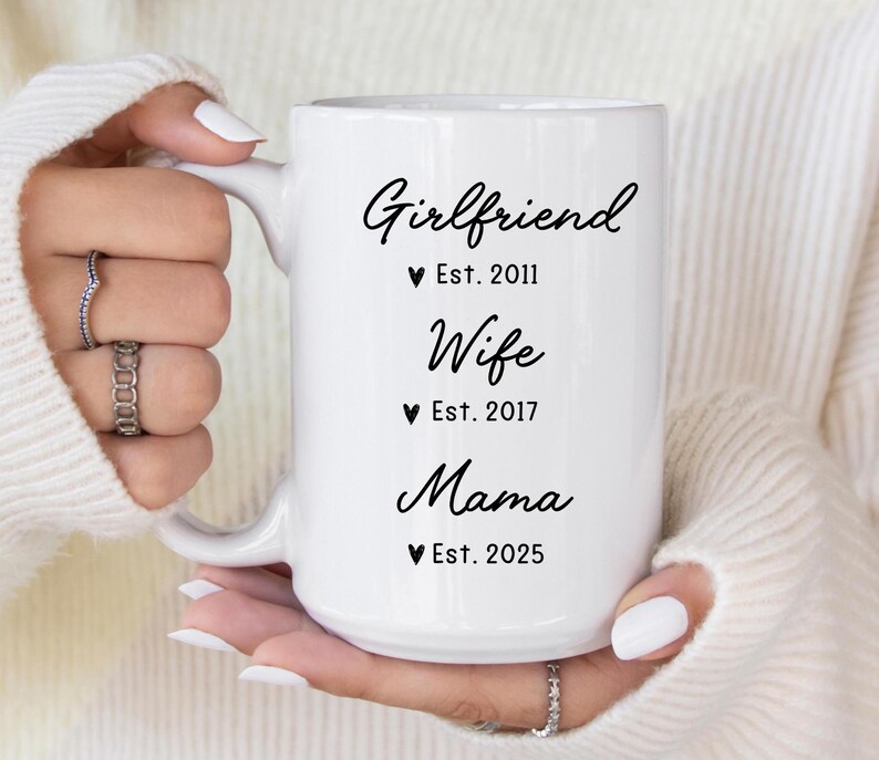 May include: White ceramic mug with black text that reads "Girlfriend Est. 2011", "Wife Est. 2017", and "Mama Est. 2025".