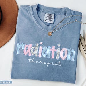 May include: A blue jean coloured t-shirt with the words "radiation therapist" in a playful font. The word "radiation" is in a mix of blue gingham and floral patterns. A gold necklace with a pendant rests on the shirt.