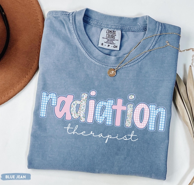 May include: A blue jean coloured t-shirt with the words "radiation therapist" in a playful font. The word "radiation" is in a mix of blue gingham and floral patterns. A gold necklace with a pendant rests on the shirt.