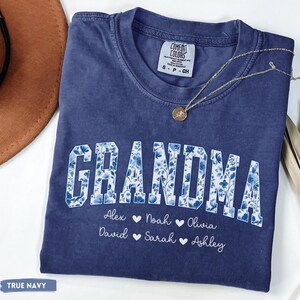 May include: A navy blue t-shirt featuring the word "GRANDMA" in white block letters with a blue floral design. Beneath the word are names: Alex, Noah, Olivia, David, Sarah, and Ashley. A gold necklace with a pendant is on the shirt.