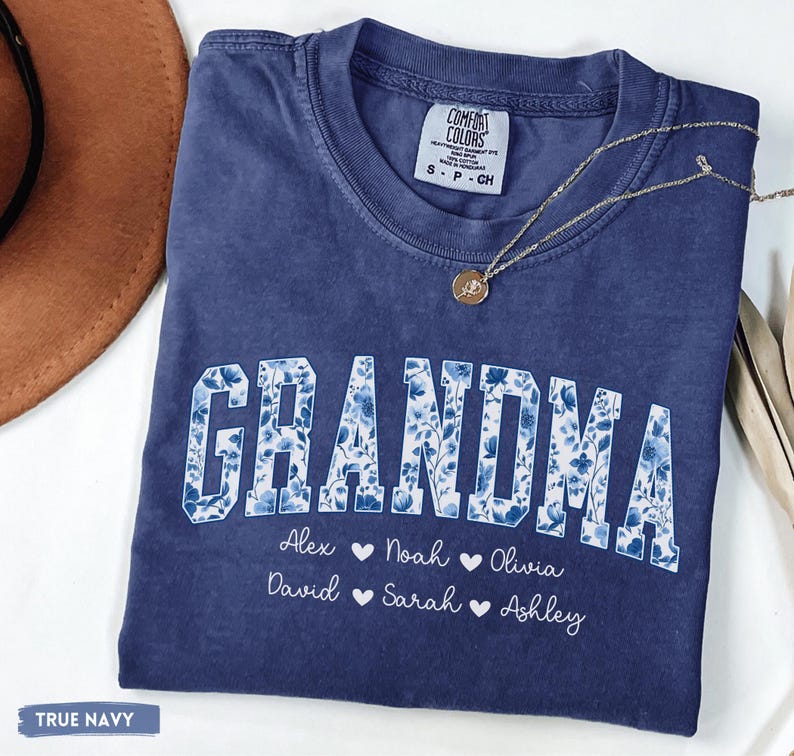 May include: A navy blue t-shirt featuring the word "GRANDMA" in white block letters with a blue floral design. Beneath the word are names: Alex, Noah, Olivia, David, Sarah, and Ashley. A gold necklace with a pendant is on the shirt.