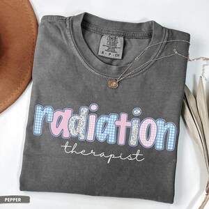 May include: A grey t-shirt featuring the text "radiation therapist". The word "radiation" is designed with a blue gingham and pink floral pattern. A gold necklace with a pendant is draped on the shirt.