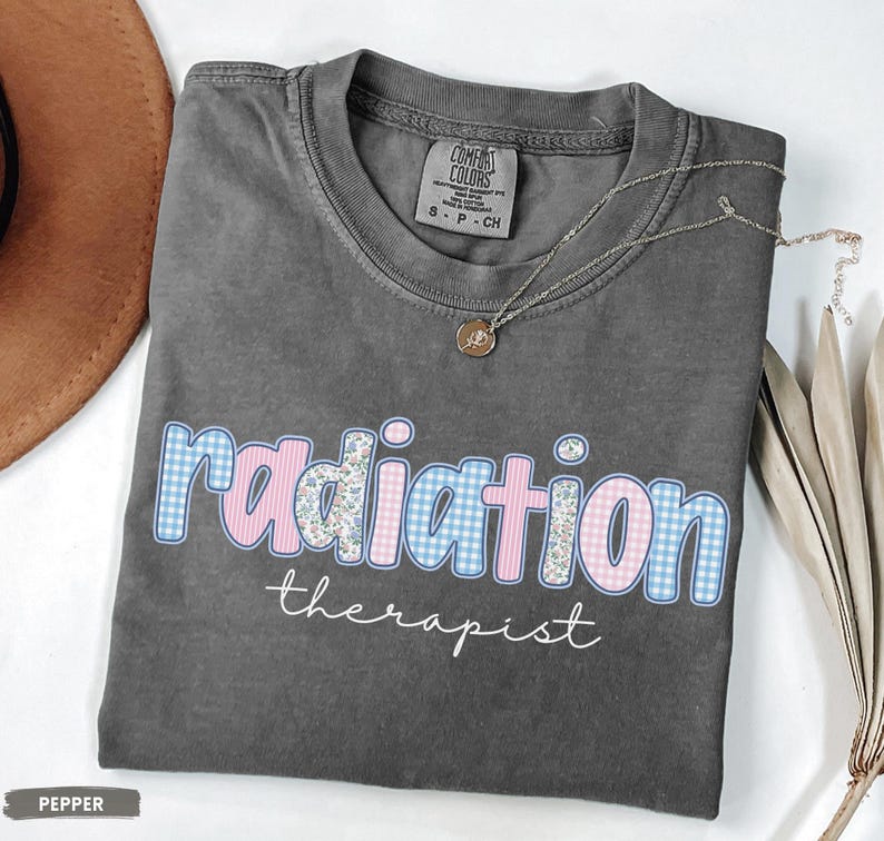 May include: A grey t-shirt featuring the text "radiation therapist". The word "radiation" is designed with a blue gingham and pink floral pattern. A gold necklace with a pendant is draped on the shirt.