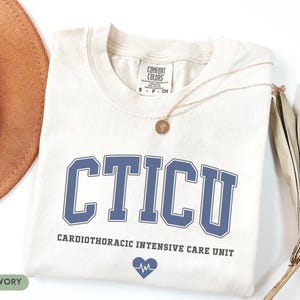May include: Ivory-colored t-shirt with the letters "CTICU" in blue, above "Cardiothoracic Intensive Care Unit" and a heart with a heartbeat graphic. A gold necklace and a brown hat are visible.