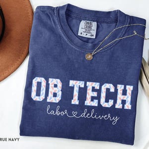 May include: A navy blue t-shirt with the words "OB TECH" in white block letters, decorated with blue and pink baby feet. The words "labor & delivery" are written below in a script font. A gold necklace with a pendant is on the shirt.