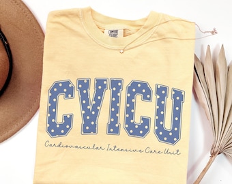 May include: A butter yellow short-sleeve t-shirt featuring the blue text CVICU in a collegiate font with white polka dots. Below the large letters, the text Cardiovascular Intensive Care Unit is printed in a smaller, cursive black font.