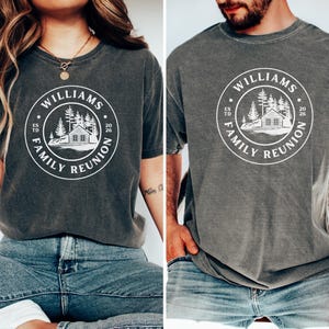 May include: A charcoal gray short-sleeve t-shirt featuring a white circular graphic on the chest. The design includes a cabin, pine trees, and the text "WILLIAMS FAMILY REUNION" with "ESTD" on the left and "2026" on the right side of the circle.