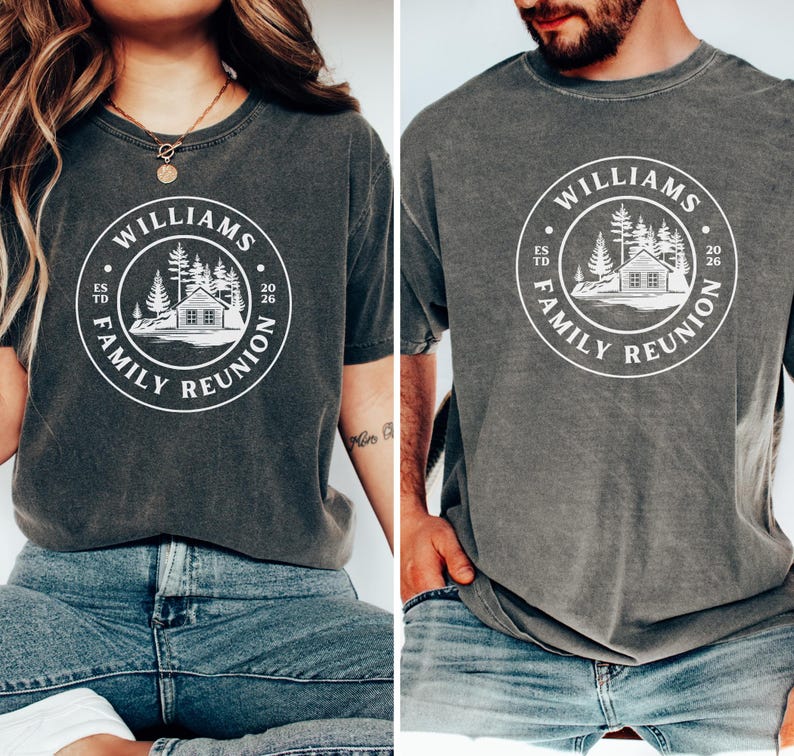 May include: A charcoal gray short-sleeve t-shirt featuring a white circular graphic on the chest. The design includes a cabin, pine trees, and the text "WILLIAMS FAMILY REUNION" with "ESTD" on the left and "2026" on the right side of the circle.