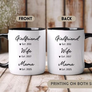 May include: White ceramic mug with a black handle and black rim. The mug has black text that reads "Girlfriend Est. 2011 Wife Est. 2017 Mama Est. 2025". The mug is printed on both sides.
