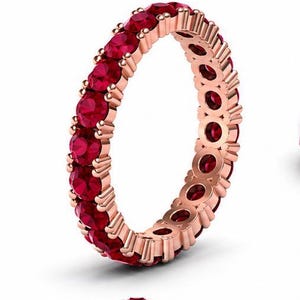 May include: An eternity band ring crafted from rose gold, featuring a continuous row of round-cut, deep red rubies set in a prong setting. The design showcases a uniform arrangement of gemstones encircling the entire circumference of the metal band.