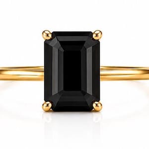 May include: A gold ring featuring a large, rectangular, emerald-cut black gemstone set in a four-prong setting. The band is thin, smooth, and polished gold, providing a clean and simple contrast to the dark, faceted stone at the center.