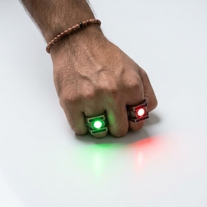 Puede incluir: Dos anillos metálicos usados en los dedos, uno con un centro circular que emite una luz verde brillante y el otro con un centro circular que emite una luz roja brillante. Ambos anillos tienen un diseño de marco rectangular y emiten luz.