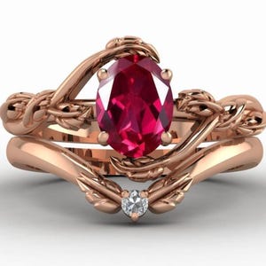 Op de afbeelding: Een roségouden verlovingsringset met een ovaal geslepen robijn en een diamantaccent. De ring heeft een op de natuur geïnspireerd ontwerp met verweven banden en blad details. De robijn is felrood.