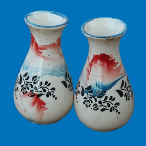 Puede incluir: Dos jarrones de cerámica blanca con borde azul y diseño floral negro. Los jarrones tienen acentos rojos y azules tipo acuarela. El fondo es azul liso.