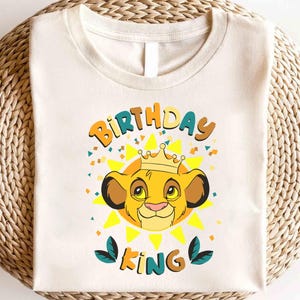 May include: Off-white t-shirt featuring a cartoon lion with a crown, surrounded by a yellow sunburst design. The text "BIRTHDAY KING" is printed above and below the lion. The shirt is on a woven surface.
