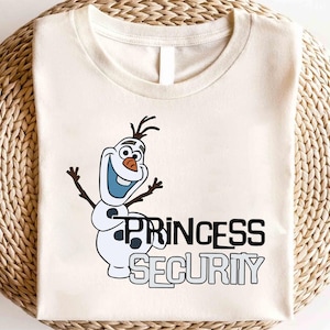 以下が含まれることがあります： クリーム色のTシャツ。雪だるまのオラフの漫画の画像と「PRINCESS SECURITY」の文字が黒とグレーで描かれています。デザインには、オラフの笑顔、腕、体が含まれています。
