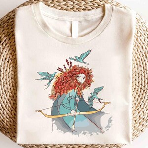 May include: A white t-shirt with a colorful illustration of a red-haired woman with a bow and arrow, surrounded by blue birds. The woman is wearing a blue dress and has a determined expression on her face.