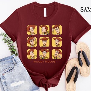 以下が含まれることがあります： トイストーリーのウッディのグラフィックがプリントされたマルーンのTシャツ。グラフィックは、ウッディの顔を9つの異なる表情で示しています。グラフィックの下には「ウッディムード」というテキストがプリントされています。