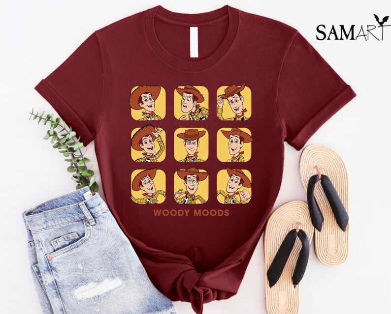 以下が含まれることがあります： トイストーリーのウッディのグラフィックがプリントされたマルーンのTシャツ。グラフィックは、ウッディの顔を9つの異なる表情で示しています。グラフィックの下には「ウッディムード」というテキストがプリントされています。