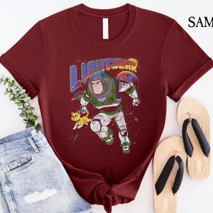 May include: A maroon t-shirt featuring a vintage-style graphic of Buzz Lightyear from Toy Story. The graphic depicts Buzz in his space ranger suit, flying through space with a small alien creature beside him. The text "LIGHTYEAR" is above Buzz in a colourful, retro font.