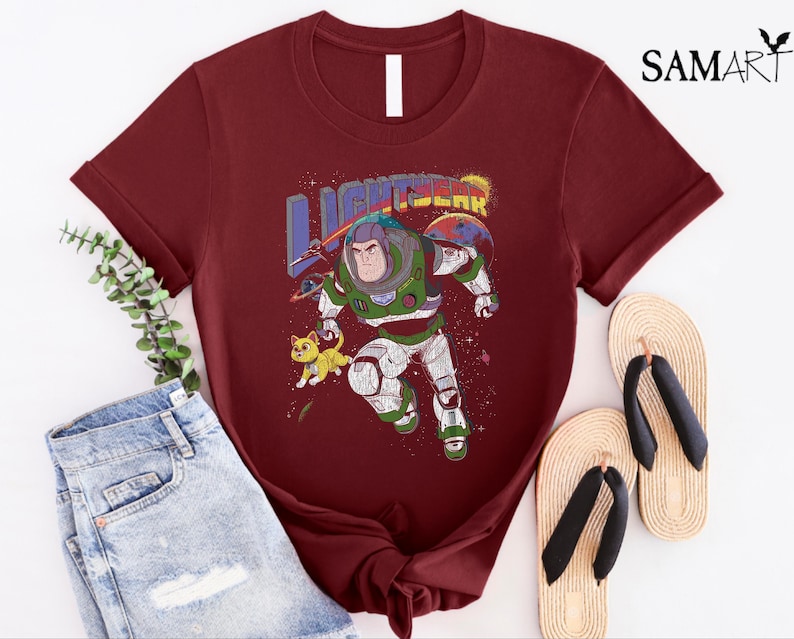 May include: A maroon t-shirt featuring a vintage-style graphic of Buzz Lightyear from Toy Story. The graphic depicts Buzz in his space ranger suit, flying through space with a small alien creature beside him. The text "LIGHTYEAR" is above Buzz in a colourful, retro font.