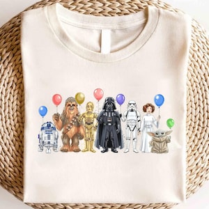 Op de afbeelding: Cr&egrave;mekleurig T-shirt met een kleurrijke illustratie van Star Wars-personages die ballonnen vasthouden. De personages zijn R2-D2, Chewbacca, C-3PO, Darth Vader, een Stormtrooper, Princess Leia en Grogu.