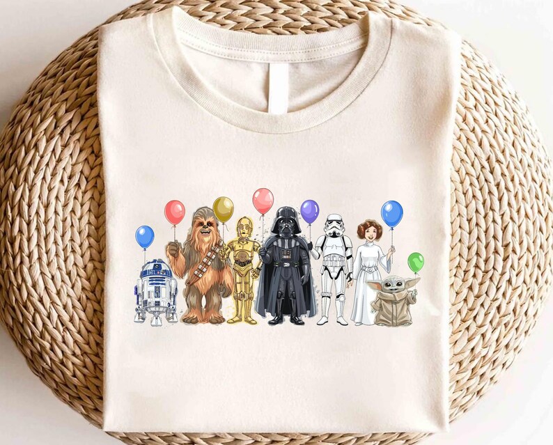 Op de afbeelding: Cr&egrave;mekleurig T-shirt met een kleurrijke illustratie van Star Wars-personages die ballonnen vasthouden. De personages zijn R2-D2, Chewbacca, C-3PO, Darth Vader, een Stormtrooper, Princess Leia en Grogu.