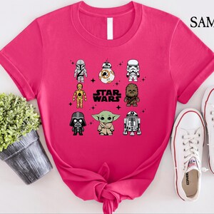 K&ouml;nnte beinhalten: Ein leuchtend pinkfarbenes T-Shirt mit einem Star Wars-Design, das Cartoon-Charaktere wie Darth Vader und Yoda zeigt. Der Text "STAR WARS" ist in Wei&szlig; gedruckt. Wei&szlig;e Turnschuhe liegen neben dem Shirt.