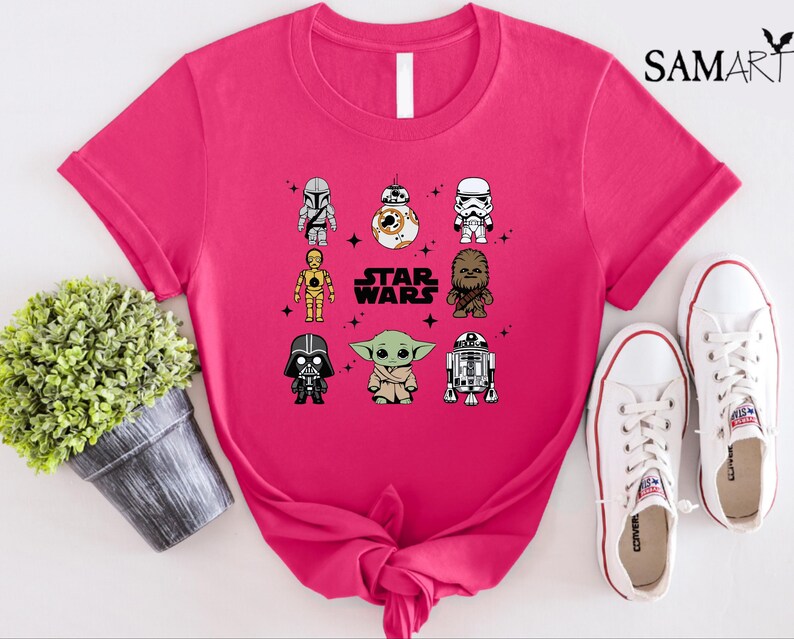 K&ouml;nnte beinhalten: Ein leuchtend pinkfarbenes T-Shirt mit einem Star Wars-Design, das Cartoon-Charaktere wie Darth Vader und Yoda zeigt. Der Text "STAR WARS" ist in Wei&szlig; gedruckt. Wei&szlig;e Turnschuhe liegen neben dem Shirt.