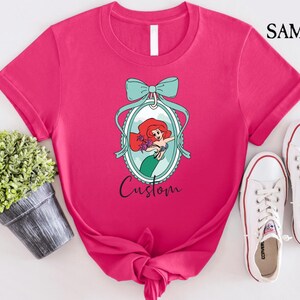 May include: A hot pink t-shirt featuring a cartoon illustration of a mermaid inside an oval frame with a teal bow. The word "Custom" is written below the frame. White sneakers are next to the shirt.