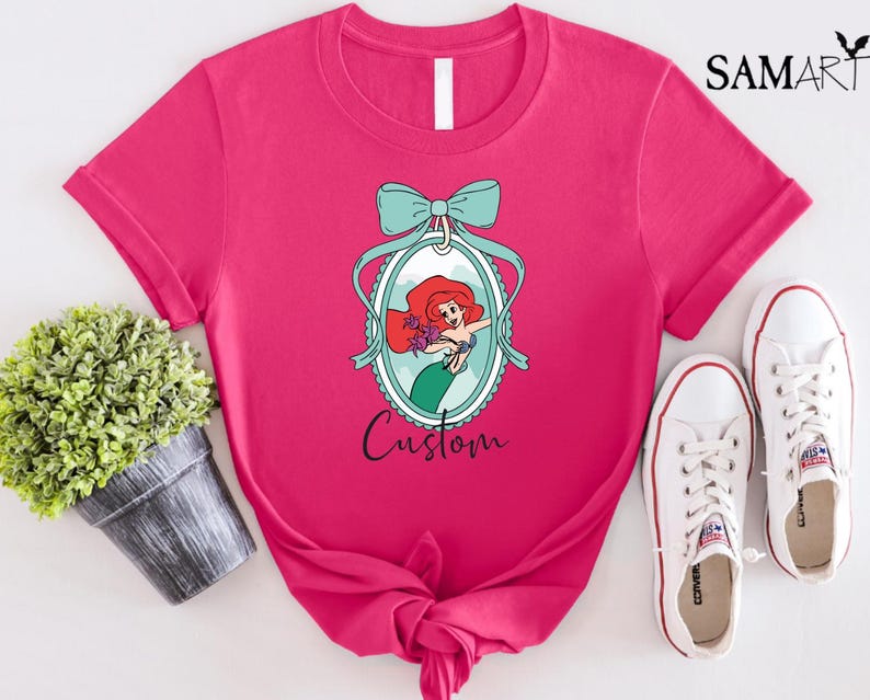 May include: A hot pink t-shirt featuring a cartoon illustration of a mermaid inside an oval frame with a teal bow. The word "Custom" is written below the frame. White sneakers are next to the shirt.