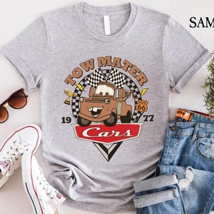 May include: A gray t-shirt with a vintage-style graphic of Tow Mater from the Disney Pixar movie Cars. The graphic features Tow Mater driving down Route 66 with the text "Tow Mater Cars" and the numbers "19" and "77".