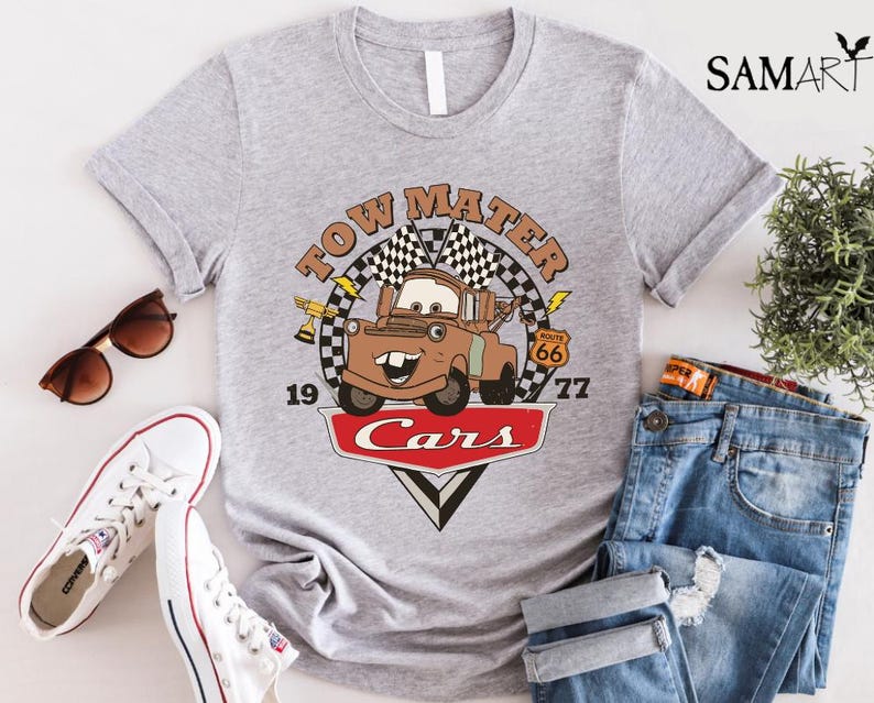 May include: A gray t-shirt with a vintage-style graphic of Tow Mater from the Disney Pixar movie Cars. The graphic features Tow Mater driving down Route 66 with the text "Tow Mater Cars" and the numbers "19" and "77".