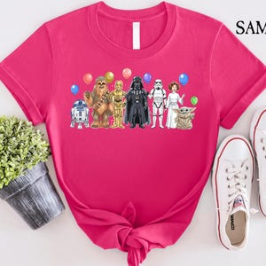 Op de afbeelding: Een felroze t-shirt met een afbeelding van Star Wars-personages en ballonnen. Het ontwerp bevat R2-D2, Chewbacca, C-3PO, Darth Vader, een Stormtrooper, prinses Leia en Grogu. Witte sneakers staan rechts.