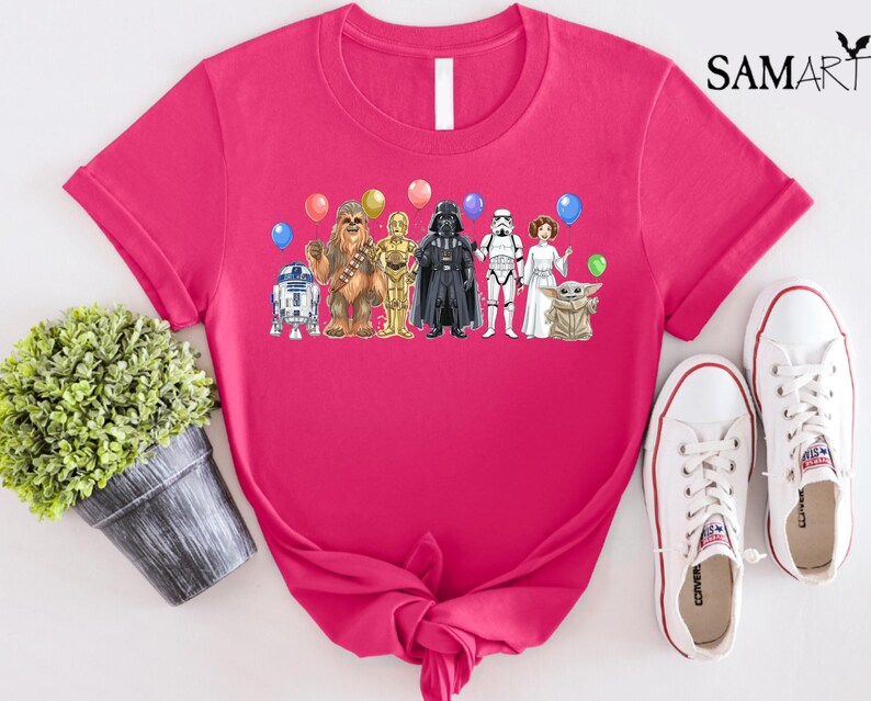 Op de afbeelding: Een felroze t-shirt met een afbeelding van Star Wars-personages en ballonnen. Het ontwerp bevat R2-D2, Chewbacca, C-3PO, Darth Vader, een Stormtrooper, prinses Leia en Grogu. Witte sneakers staan rechts.