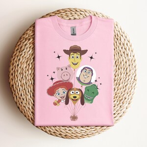 Op de afbeelding: Een lichtroze t-shirt met een afbeelding van stripfiguren gerangschikt als ballonnen. De karakters zijn onder meer een cowboy, een varken, een ruimteranger, een cowgirl, een hond en een dinosaurus. Zwarte steraccenten zijn ook aanwezig.