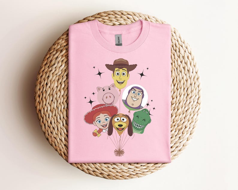 Op de afbeelding: Een lichtroze t-shirt met een afbeelding van stripfiguren gerangschikt als ballonnen. De karakters zijn onder meer een cowboy, een varken, een ruimteranger, een cowgirl, een hond en een dinosaurus. Zwarte steraccenten zijn ook aanwezig.
