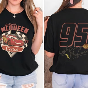 Op de afbeelding: Zwart T-shirt met een vintage-stijl afbeelding van Lightning McQueen uit de Disney Pixar film Cars. De afbeelding bevat de tekst "Lightning McQueen", "Cars" en de nummers "1997". De achterkant van het shirt heeft het nummer "95" in roodbruin met de tekst "Lightning McQueen" en een klein Rust-eze logo.