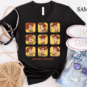 以下が含まれることがあります： トイストーリーのウッディの顔が9つ並んだ、黒いTシャツ。それぞれのウッディは異なる表情をしています。グリッドの下には「WOODY MOODS」という文字が印刷されています。
