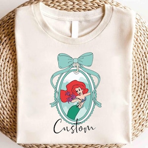 May include: Cream-colored t-shirt featuring a cartoon illustration of a mermaid with red hair and a green tail, framed by a teal bow and oval border. The word "Custom" is written in cursive below the image.