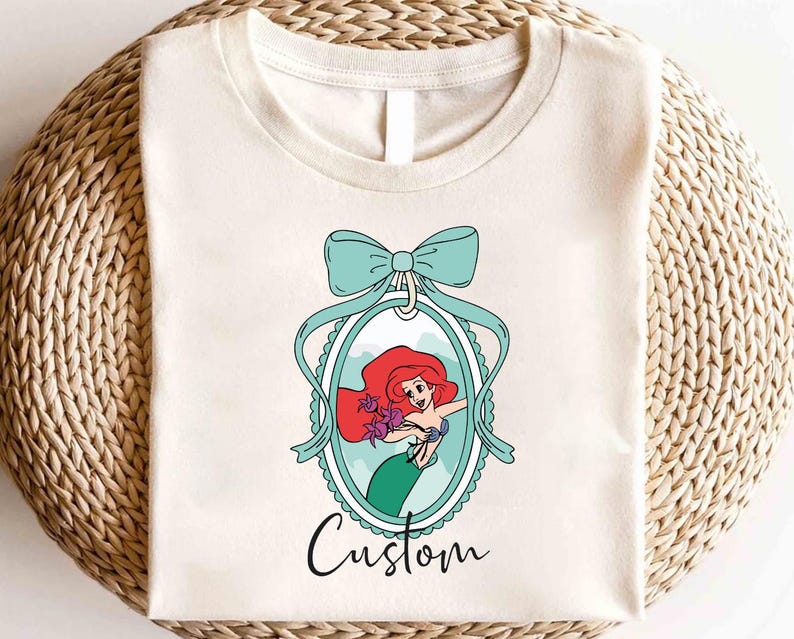 May include: Cream-colored t-shirt featuring a cartoon illustration of a mermaid with red hair and a green tail, framed by a teal bow and oval border. The word "Custom" is written in cursive below the image.