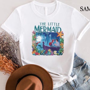 Puede incluir: Camiseta blanca con un gr&aacute;fico colorido de "La Sirenita". El dise&ntilde;o incluye el t&iacute;tulo, un barco con personajes y elementos submarinos. La camiseta est&aacute; hecha de un material suave y tiene mangas cortas.