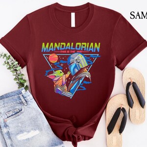 May include: A maroon t-shirt with a graphic of a Mandalorian and Grogu. The graphic is in a retro style with a triangle and the text "Mandalorian - This is the Way".