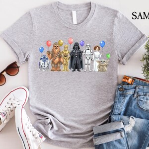 Op de afbeelding: Een lichtgrijs t-shirt met een kleurrijke afbeelding van Star Wars-personages die ballonnen vasthouden. De personages zijn onder meer R2-D2, Chewbacca, C-3PO, Darth Vader, een Stormtrooper, prinses Leia en Grogu. Het shirt wordt gecombineerd met een blauwe jeans en witte sneakers.